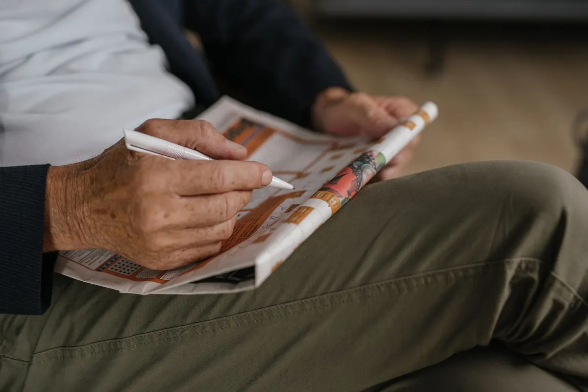 Een hand met een pen vult een kruiswoordraadsel in de krant in.